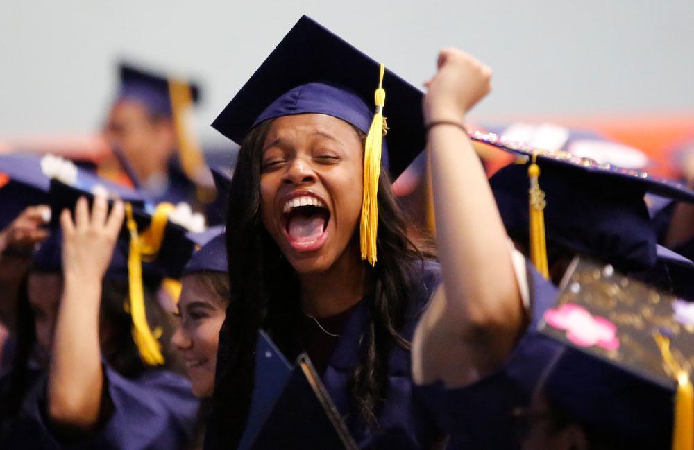 A woman in a graduation gown joyfully celebrates her achievement with a big smile and raised arms.